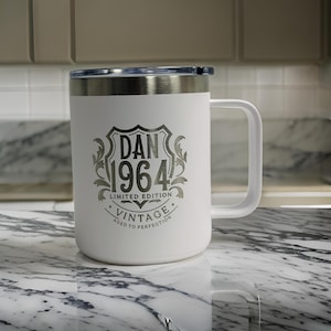 Puede incluir: Una taza blanca de acero inoxidable con asa y borde met&aacute;lico. El frente presenta un dise&ntilde;o de escudo grabado en gris oscuro con el texto DAN 1964 LIMITED EDITION VINTAGE AGED TO PERFECTION, centrado dentro de un motivo de hojas.