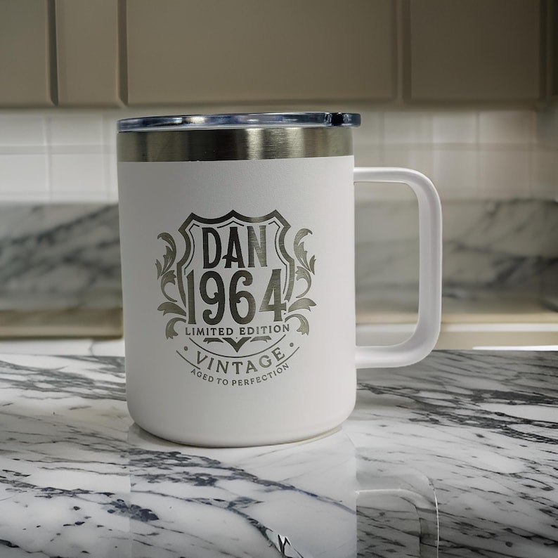 Puede incluir: Una taza blanca de acero inoxidable con asa y borde met&aacute;lico. El frente presenta un dise&ntilde;o de escudo grabado en gris oscuro con el texto DAN 1964 LIMITED EDITION VINTAGE AGED TO PERFECTION, centrado dentro de un motivo de hojas.