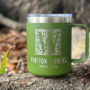 Puede incluir: Una taza t&eacute;rmica de color verde oliva mate con asa y tapa transparente. El frente presenta una letra W grande rodeada por un patr&oacute;n de hojas, con el texto WINSTON FLOWERS. since 1944 grabado en color plata claro sobre la superficie.