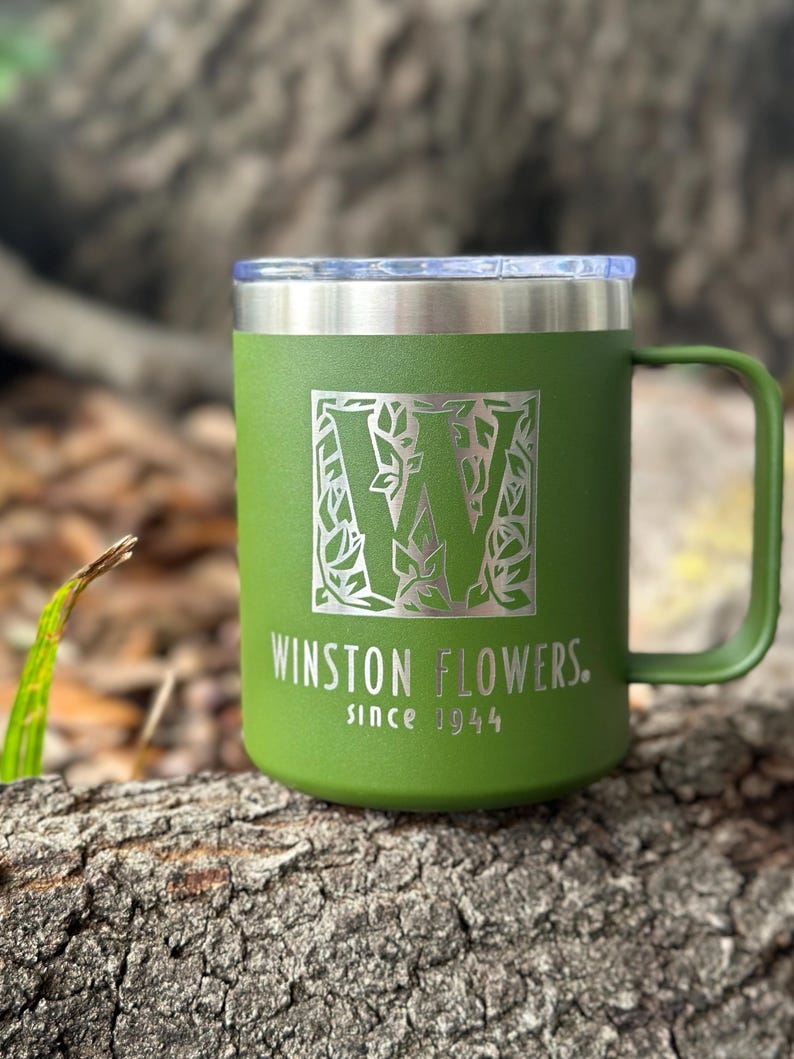 Puede incluir: Una taza t&eacute;rmica de color verde oliva mate con asa y tapa transparente. El frente presenta una letra W grande rodeada por un patr&oacute;n de hojas, con el texto WINSTON FLOWERS. since 1944 grabado en color plata claro sobre la superficie.