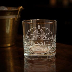 May include: A clear glass tumbler featuring an etched design of a bottle centered within a semi-circular frame. The text THE VAULT is engraved below the bottle, with EST 2025 positioned underneath. Small card suit symbols flank the central graphic.