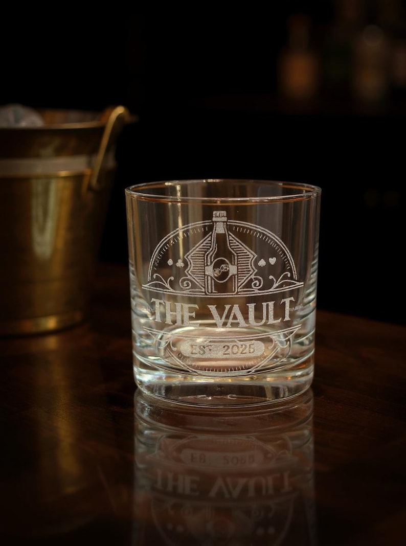 May include: A clear glass tumbler featuring an etched design of a bottle centered within a semi-circular frame. The text THE VAULT is engraved below the bottle, with EST 2025 positioned underneath. Small card suit symbols flank the central graphic.