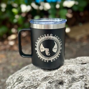Puede incluir: Una taza de acero inoxidable negra con tapa transparente y asa. El frente presenta un logotipo circular con el texto "FRANCISKO JAVYER VINTAGE BARBER" y "EST. 2025" rodeando la silueta de una persona que lleva una gorra plana en el centro.