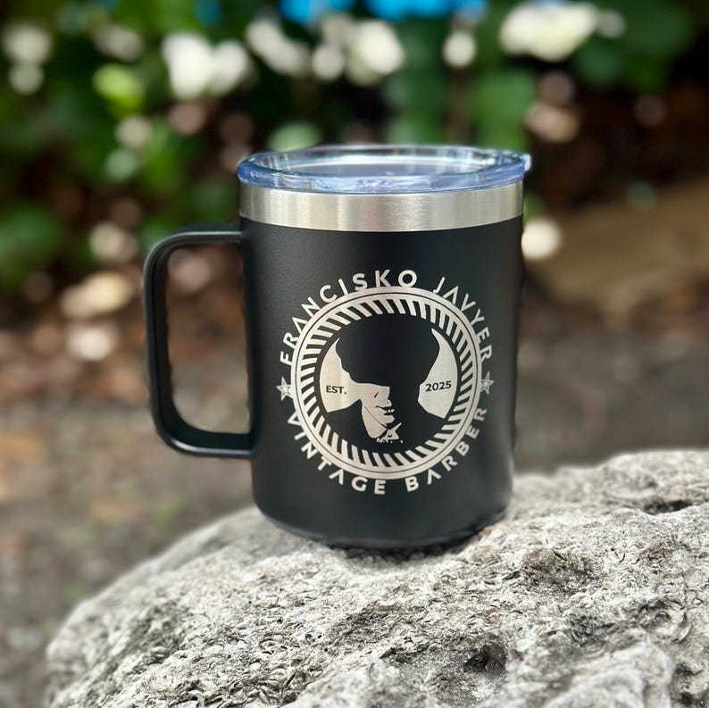 Puede incluir: Una taza de acero inoxidable negra con tapa transparente y asa. El frente presenta un logotipo circular con el texto "FRANCISKO JAVYER VINTAGE BARBER" y "EST. 2025" rodeando la silueta de una persona que lleva una gorra plana en el centro.