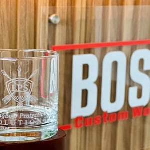 May include: A clear glass tumbler featuring an etched design of a shield with crossed arrows and the letters LPS. Below the shield, the text reads LongBo Protection SOLUTIONS. The glass has a thick base and straight sides, showing clear glass.