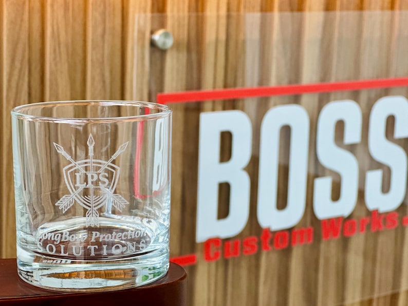 May include: A clear glass tumbler featuring an etched design of a shield with crossed arrows and the letters LPS. Below the shield, the text reads LongBo Protection SOLUTIONS. The glass has a thick base and straight sides, showing clear glass.