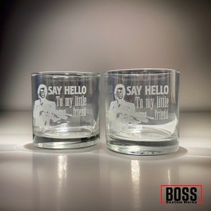 May include: Two clear glass tumblers featuring an etched design of a person holding a firearm with the text SAY HELLO To my little friend. The glasses have a thick base and straight sides, with a BOSS Custom Works logo in the bottom right corner.