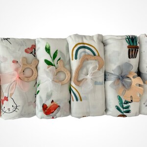 May include: Five white muslin swaddles with different colorful patterns, each tied with a ribbon and a wooden teether. The patterns include floral, rainbow, and animal prints.