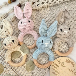 May include: Four crocheted bunny rattle toys in pink, blue, beige, and brown with wooden rings. The toys are on a beige crocheted blanket with a wooden sign that says "hello world" and has spaces for name, date, weight, and length.