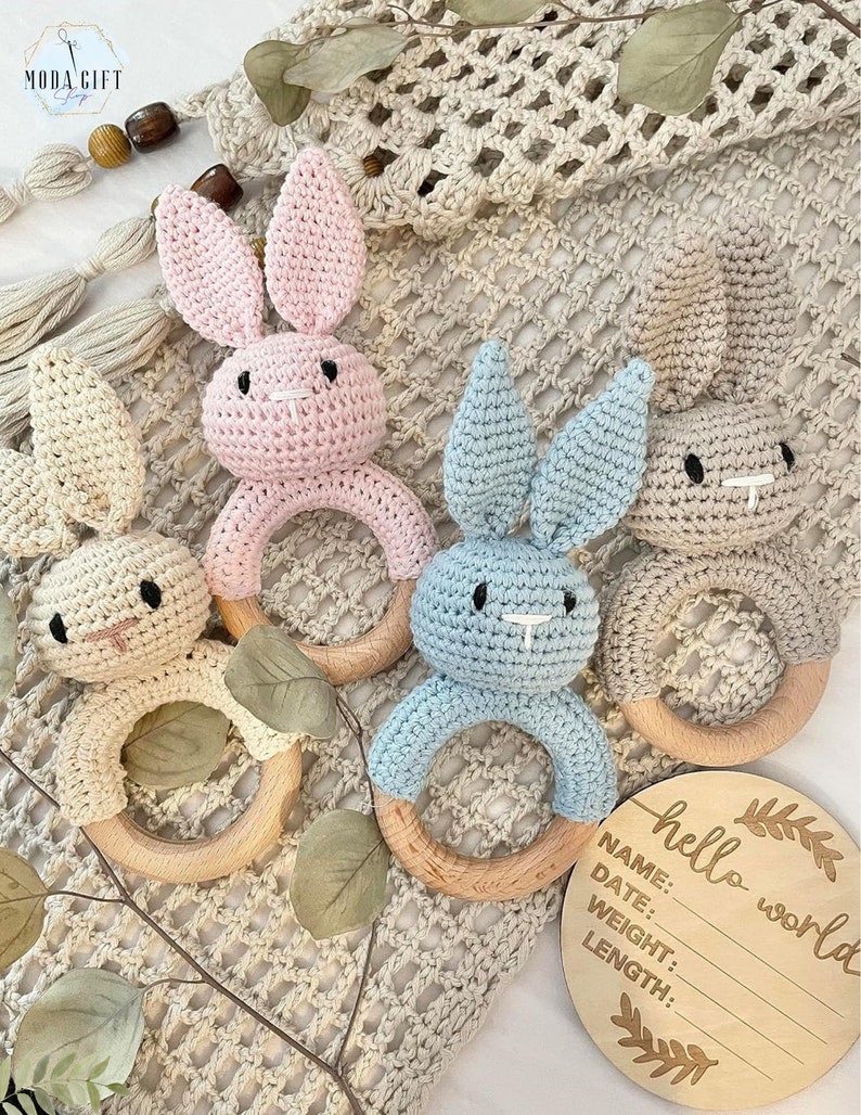Pu&ograve; includere: Quattro giocattoli sonagli a forma di coniglio all'uncinetto in rosa, blu, beige e marrone con anelli di legno. I giocattoli sono su una coperta di uncinetto beige con un cartello di legno che dice "hello world" e ha spazi per il nome, la data, il peso e la lunghezza.
