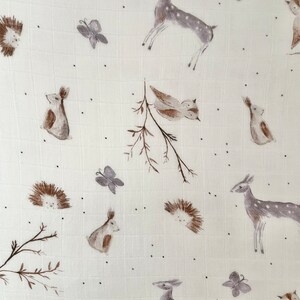 May include: A white muslin swaddle blanket with a woodland animal print. The print features deer, birds, butterflies, hedgehogs, and rabbits in shades of brown and gray.