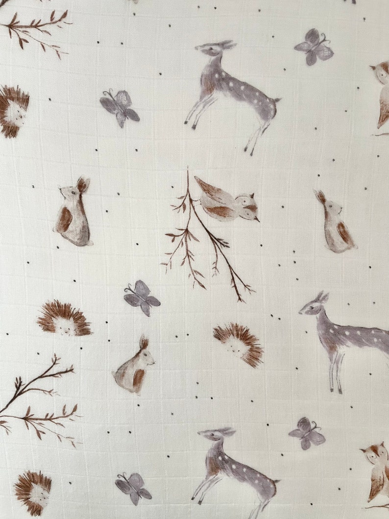 May include: A white muslin swaddle blanket with a woodland animal print. The print features deer, birds, butterflies, hedgehogs, and rabbits in shades of brown and gray.