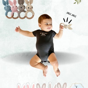 Pu&ograve; includere: Un set di quattro sonagli a forma di coniglio all'uncinetto con anelli di legno. I giocattoli sono in blu, rosa, grigio e beige. Un bambino tiene un sonaglio simile. Il testo "Jingle Jingle" &egrave; scritto sopra il bambino. Il testo "HANDSOME LITTLE FELLA" &egrave; sulla maglietta del bambino. L'immagine include anche una fila di cinque coniglietti dei cartoni animati in basso.
