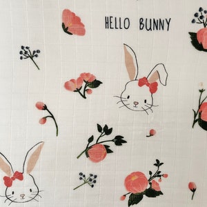 May include: White muslin baby blanket with a pink and green floral pattern and bunny illustrations. The text "Hello Bunny" is repeated twice on the blanket.