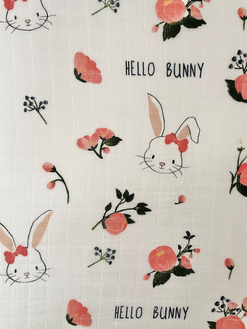 May include: White muslin baby blanket with a pink and green floral pattern and bunny illustrations. The text "Hello Bunny" is repeated twice on the blanket.