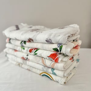May include: A stack of eight white muslin baby swaddles with different colorful floral and animal prints.