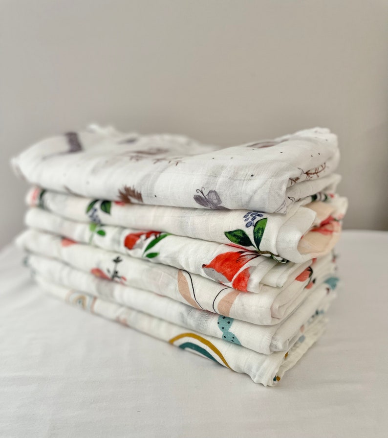 May include: A stack of eight white muslin baby swaddles with different colorful floral and animal prints.