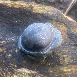 May include: A smooth, dark gray, round glass bottle stopper with a slight indentation on the top.