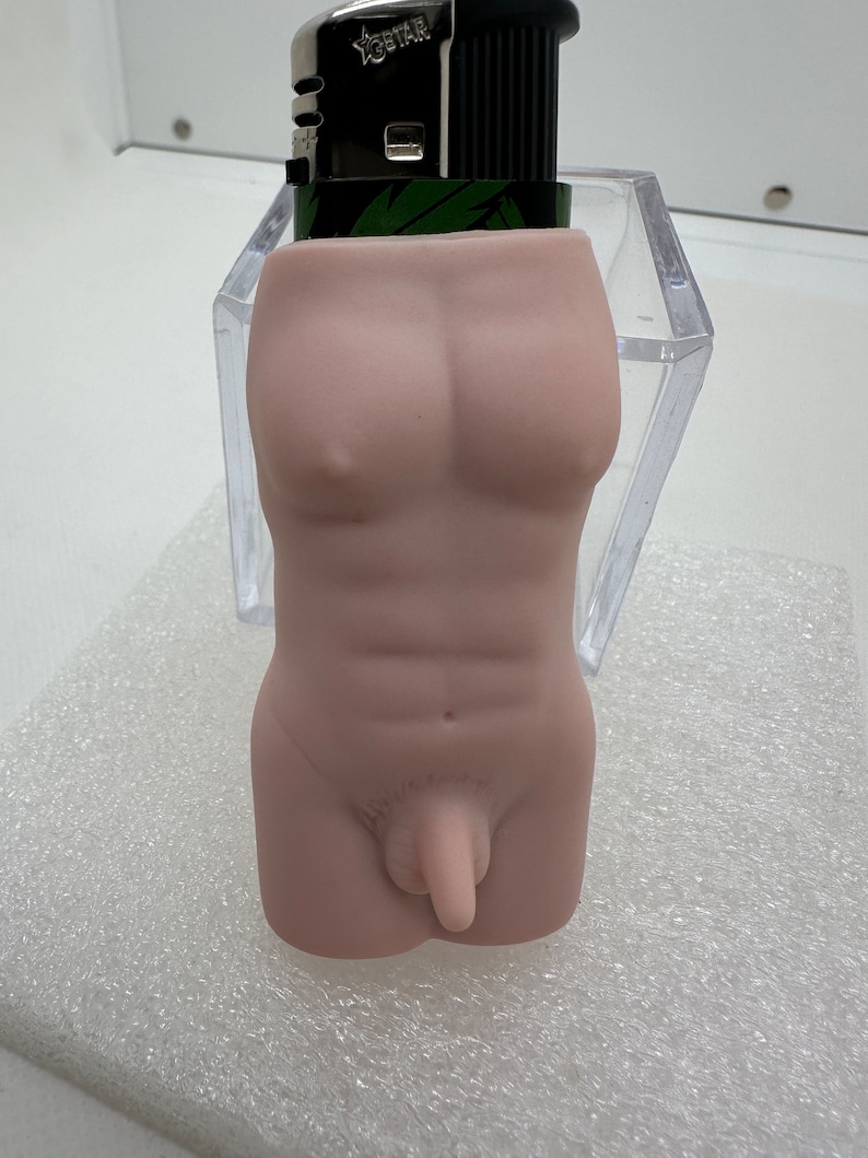 May include: A nude-colored lighter holder shaped like a human torso. The lighter holder is made of a smooth, matte material. The lighter holder is sitting on a white, textured surface.
