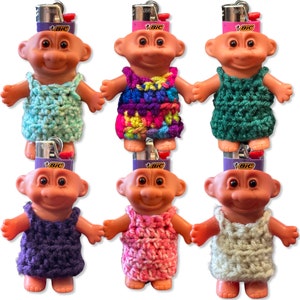 May include: Six troll dolls with different colored crocheted dresses. Each doll is wearing a dress made of a different color yarn. The dolls are all wearing a Bic lighter on their head.