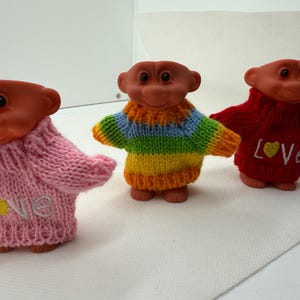 May include: Three vintage troll dolls with orange skin and knitted sweaters. One doll wears a pink sweater with "Love" embroidered on it, another wears a rainbow striped sweater, and the third wears a red sweater with "Love" embroidered.