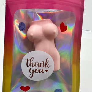 May include: A pink,  silicone bust shaped like a woman's torso with a white circle sticker that says "thank you" with a small heart. The bust is in a clear plastic bag with a holographic rainbow pattern. The bag is sealed with a pink zipper.