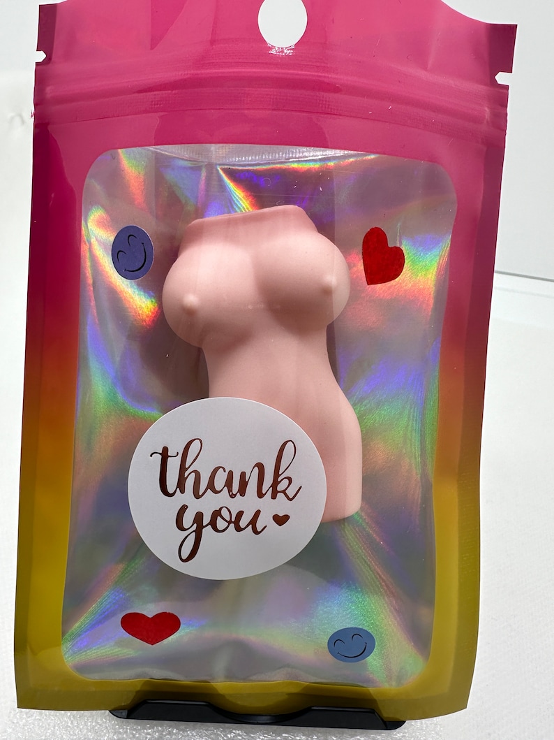 May include: A pink,  silicone bust shaped like a woman's torso with a white circle sticker that says "thank you" with a small heart. The bust is in a clear plastic bag with a holographic rainbow pattern. The bag is sealed with a pink zipper.