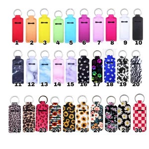 May include: A collection of thirty rectangular neoprene lip balm holders with metal key rings, featuring various solid colors, marble patterns, animal prints, floral designs, and geometric motifs, each numbered from one to thirty for identification.