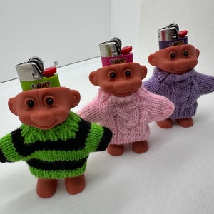 May include: Three small troll dolls wearing knitted sweaters. The dolls have red hair and are wearing a green, pink, and purple sweater. Each doll has a Bic lighter on their head.