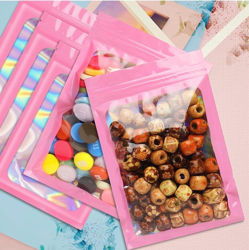 May include: Several pink, resealable bags with clear windows. One bag contains colorful candies, while others hold wooden beads in various shades of brown and tan. The bags have a holographic, iridescent strip on the side.