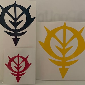 May include: Three vinyl stickers featuring a stylized emblem in black, red, and yellow. The emblem has a central leaf-like design within a circular frame with pointed extensions. The stickers vary in size, with the largest in black.