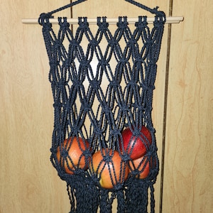 May include: A dark blue macrame wall hanging with a wooden dowel at the top. The hanging has a fringe at the bottom and is filled with red and orange apples.