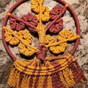 May include: A macrame wall hanging with a tree design. The tree is made of brown and yellow rope and has a fringe border.