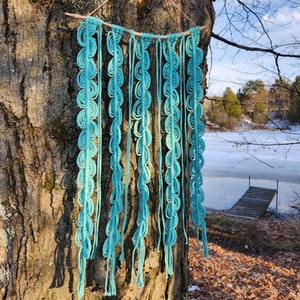 May include: A turquoise macrame wall hanging with a wooden dowel at the top. The hanging is made of multiple strands of yarn that are knotted together to create a series of loops. The hanging is hanging from a tree branch.