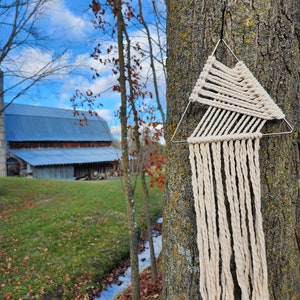 May include: A white macrame wall hanging with a triangular frame, hanging from a tree trunk. The macrame is made with thick white rope and has a fringe at the bottom.