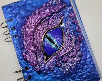 May include: A blue spiral bound notebook with a purple and blue glitter dragon eye on the cover.