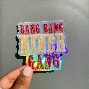 May include: A holographic sticker with the text "Bang Bang Niner Gang" in red and beige lettering.
