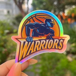 May include: A holographic sticker featuring the Golden State Warriors logo. The logo is a blue silhouette of a basketball player with a lightning bolt, set against an orange basketball with a net. The text "WARRIORS" is in yellow and orange.