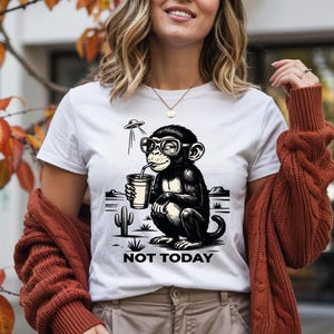 May include: White t-shirt featuring a black and white illustration of a monkey wearing glasses and drinking from a cup. The design includes a UFO and the text "NOT TODAY". The monkey is set against a desert landscape.