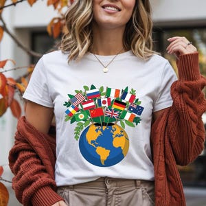 Könnte beinhalten: Weißes T-Shirt mit einer Grafik eines blauen und gelben Globus, aus dem die Flaggen verschiedener Nationen herausragen. Die Flaggen repräsentieren verschiedene Länder und symbolisieren globale Einheit und Vielfalt. Das T-Shirt ist ein lässiges Kleidungsstück für den Alltag.