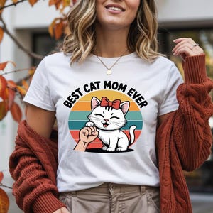May include: White t-shirt with the text "BEST CAT MOM EVER" in black. The graphic features a cartoon cat with a red bow, a hand, and a colorful sunset design. The shirt is a casual wear item.
