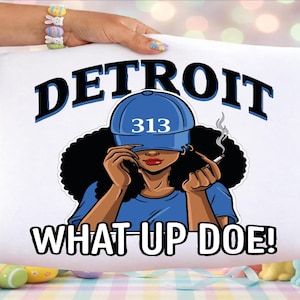 Puede incluir: Cojín blanco con un gráfico de una persona con una gorra azul con el número "313" y la palabra "DETROIT" encima. La frase "WHAT UP DOE!" está debajo.