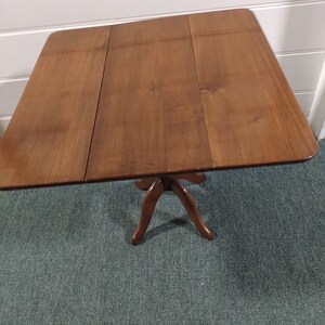 May include: A brown wooden folding table with a pedestal base. The table has a rectangular top and three legs.