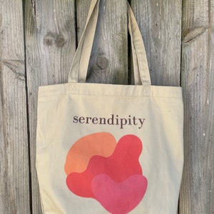 Puede incluir: Una bolsa de tela beige con la palabra "serendipity" impresa en texto marr&oacute;n. La bolsa tiene un dise&ntilde;o abstracto rojo, naranja y rosa.