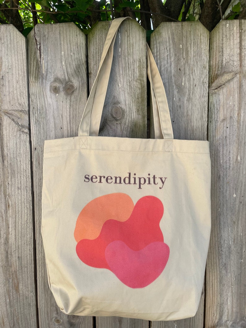Puede incluir: Una bolsa de tela beige con la palabra "serendipity" impresa en texto marr&oacute;n. La bolsa tiene un dise&ntilde;o abstracto rojo, naranja y rosa.