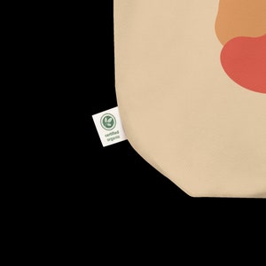 Puede incluir: Una bolsa de tela beige con una etiqueta verde certificada como org&aacute;nica. La bolsa tiene un dise&ntilde;o de formas abstractas en naranja, amarillo y rojo.