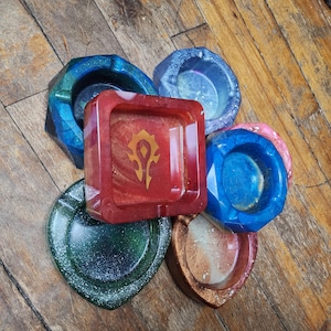 Può includere: Una collezione di posacenere fatti a mano in varie forme e colori, tra cui blu, verde, rosso e marrone. I posacenere sono in resina e hanno una finitura lucida. Un posacenere presenta un emblema dorato.