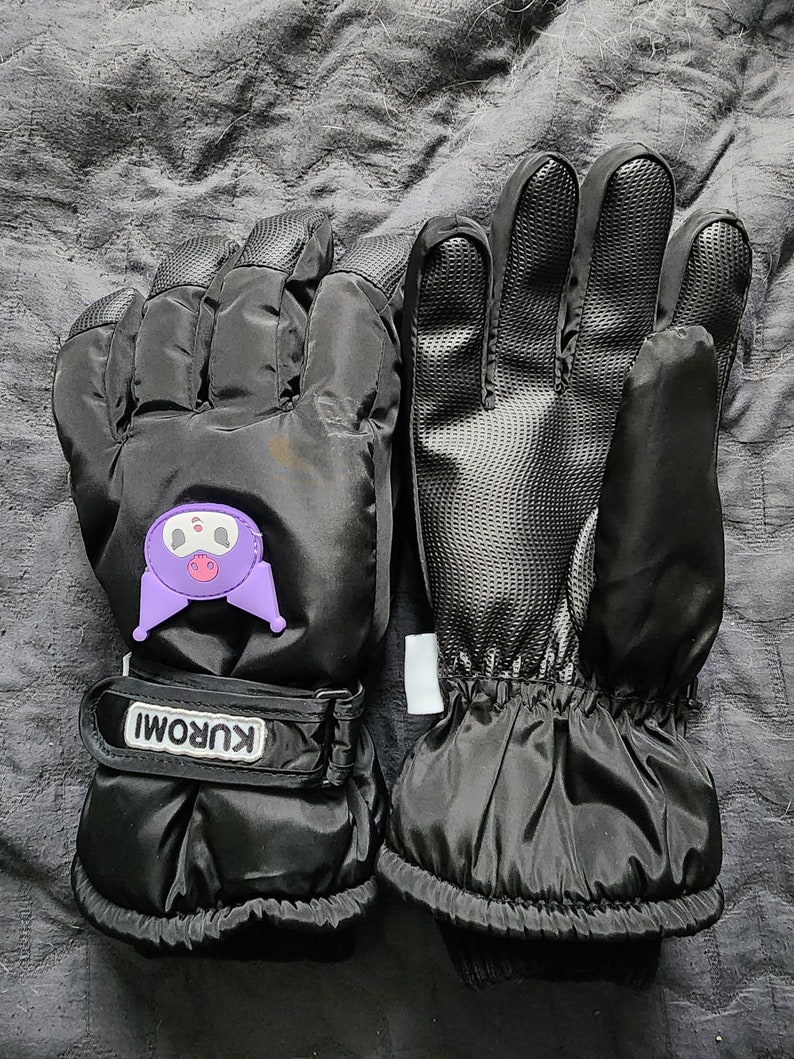 K&ouml;nnte beinhalten: Ein Paar schwarze Winterhandschuhe mit einer lilafarbenen Comicfigur auf einem Handschuh. Die Handschuhe haben einen schwarzen Riemen mit dem Wort "KUROMI" darauf.