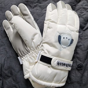 K&ouml;nnte beinhalten: Ein Paar wei&szlig;e Winterhandschuhe mit einem schwarzen Klettverschluss und dem aufgedruckten Wort "rochacco". Die Handschuhe haben ein wei&szlig;es, flauschiges Futter und ein kleines wei&szlig;es Comic-Tier auf der R&uuml;ckseite eines Handschuhs.