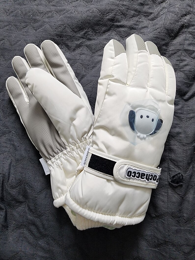 K&ouml;nnte beinhalten: Ein Paar wei&szlig;e Winterhandschuhe mit einem schwarzen Klettverschluss und dem aufgedruckten Wort "rochacco". Die Handschuhe haben ein wei&szlig;es, flauschiges Futter und ein kleines wei&szlig;es Comic-Tier auf der R&uuml;ckseite eines Handschuhs.